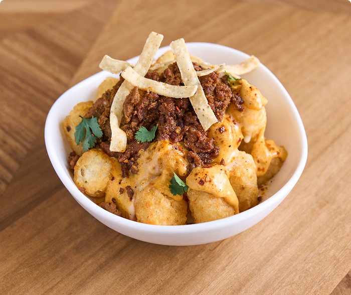 Potato tots and beef taco meat in a white bowl covered in nacho cheese, creamy sauce, fresh cilantro and tortilla strips