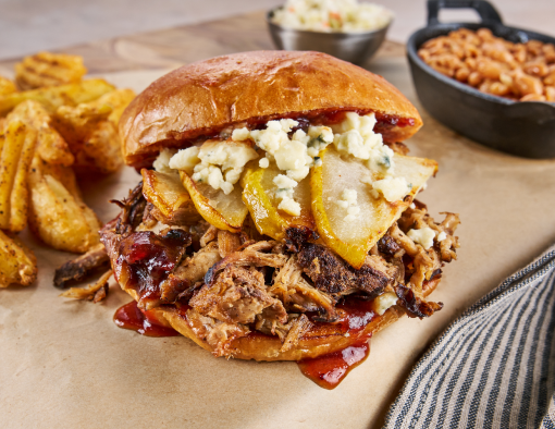 A pork barbecue sandwich stuffed with cooked pear slices and blue cheese crumbles with side dishes in the background