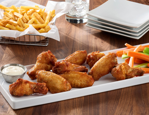 Roasted bone-in chicken wings arranged on a white platter with dipping sauce, carrot and celery sticks, and fries on a wooden table