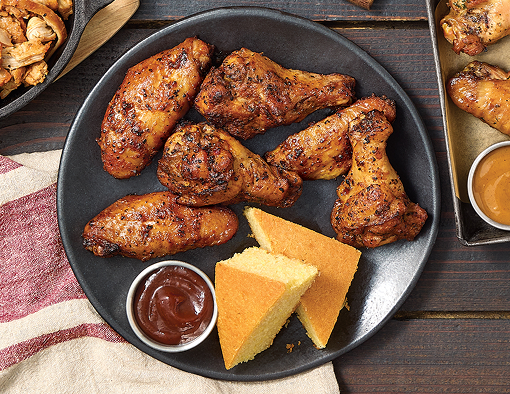 Sweet heat smoked chicken wings arranged on a black plate with cornbread wedges and a cup of barbecue sauce on a rustic wooden table