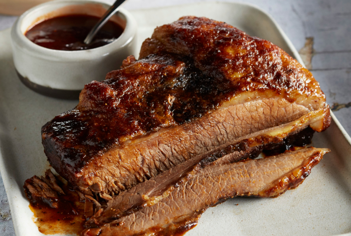 Partially sliced barbecue brisket glazed with sauce and served with additional barbecue sauce on the side.