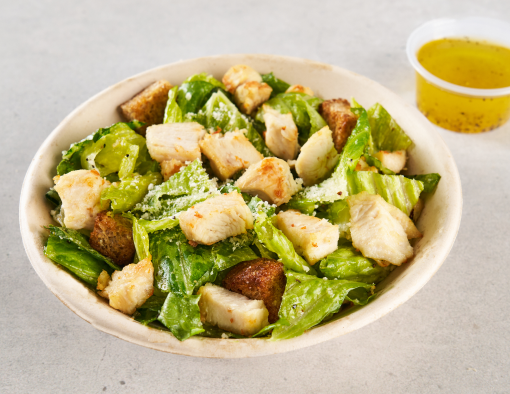 A bowl filled with romaine lettuce, chicken chunks croutons, and ramekin of dressing on the side
