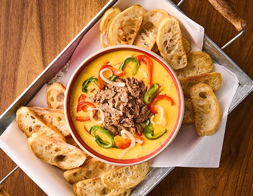 A bowl of creamy cheese dip topped with steak and peppers, served with toasted bread slices on a tray