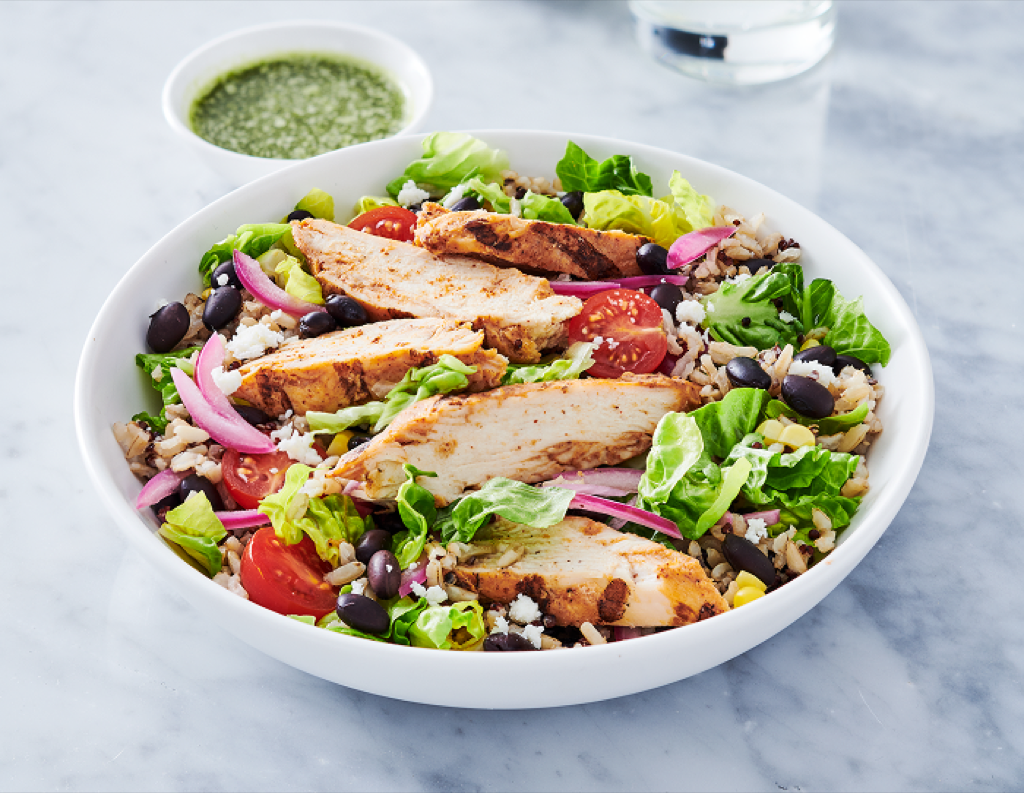 White bowl with mixed greens, rice, black beans and vegetables topped with slices of grilled chicken