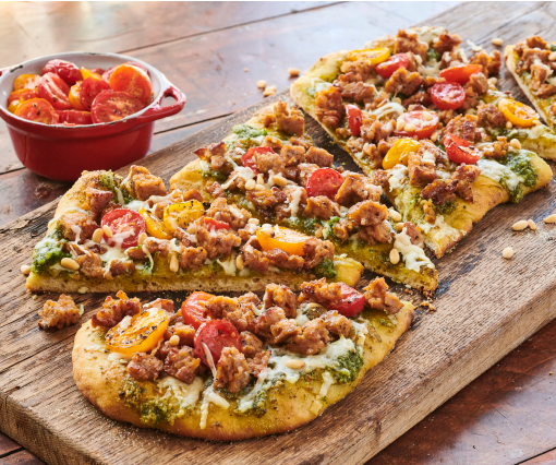 Pesto flatbread pizza sliced on a wooden board, topped with sausage crumbles, melted cheese, pesto, and red and yellow cherry tomatoes