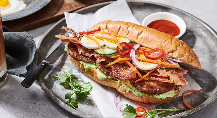 Breakfast Bánh Mì sandwich featuring sausage patties, bacon and fresh vegetables and a side of dipping sauce