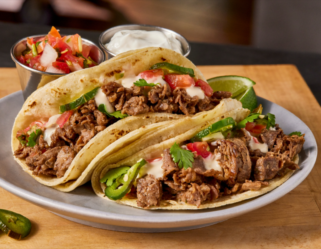 plate of two soft tacos stuffed with sliced beef, tomatoes and jalapeños with sides of Pico de Gallo and sour cream