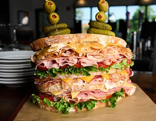 A tall, multi-layered sandwich with meats, cheeses, lettuce, tomato, with olives and pickles on skewers