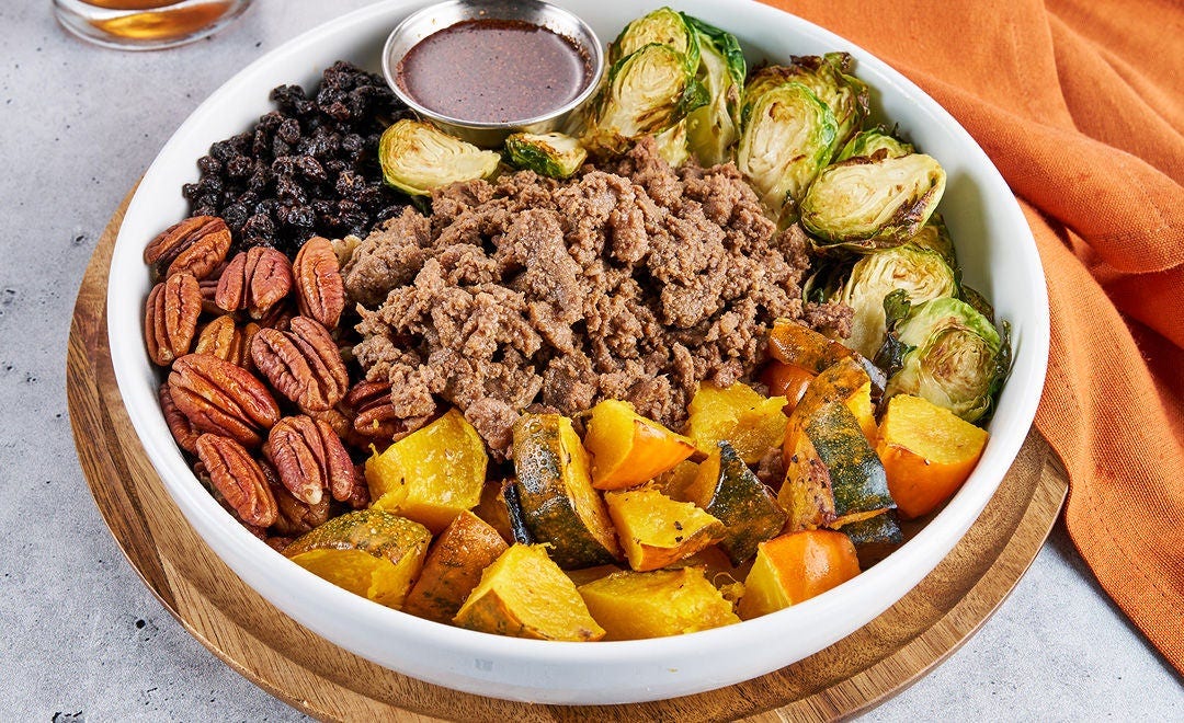 A bowl loaded with sliced beef, acorn squash, pecans, black currants, Brussels sprouts and sauce in a metal ramekin