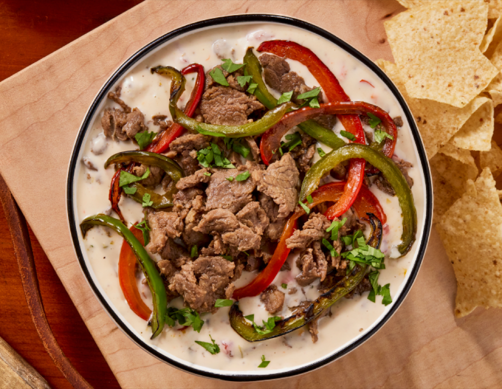 bowl of queso dip topped with sliced beef, sliced peppers, cilantro with a side of tortilla chips
