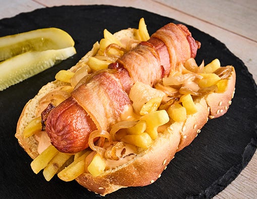 A bacon-wrapped hot dog topped with fries and grilled onions, served with pickles on a black plate