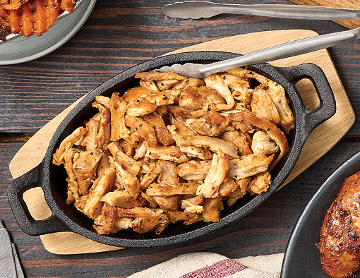 Cast-iron pan filled with sweet heat smoked chicken thigh pieces, lightly sauced and shredded, resting on a wooden board with metal tongs nearby