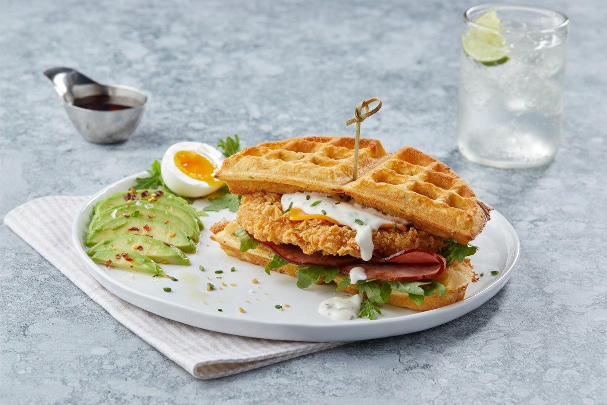 Image of chicken waffle sandwich on a plate with a side of sliced avocado and a soft boiled egg.