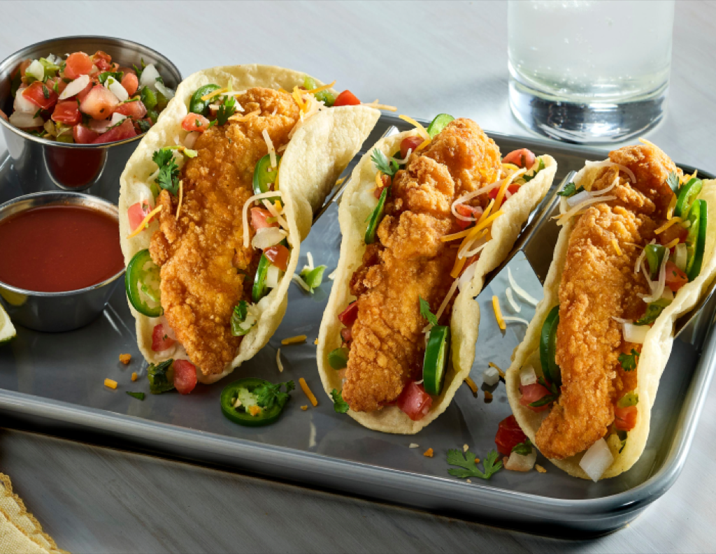 Three gluten-free breaded chicken tender tacos with lettuce, jalapeños, pico de gallo, and dipping sauces on a metal tray