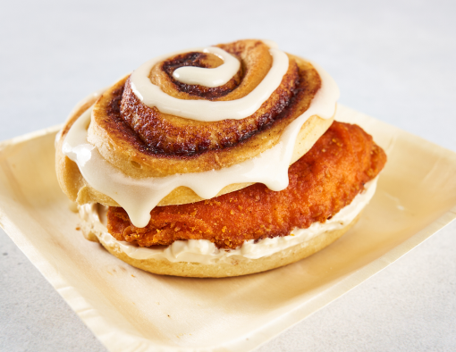 A disposable plate holding a cinnamon roll with a crispy chicken filet and maple cream sandwiched in between