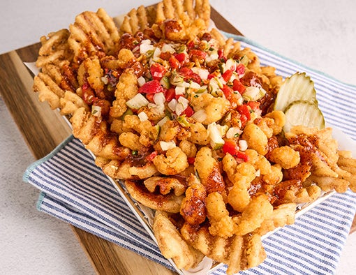 Waffle fries topped with crispy chicken bites, red sauce, relish, and pickles in a basket