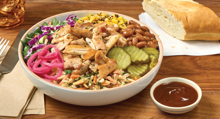 Bowl of smoked chicken grain bowl with sliced chicken, beans, pickles, pickled onions, slaw, grains, and barbecue sauce on wooden table