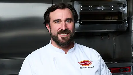 A bearded Tyson Foodservice chef smiling warmly in a restaurant kitchen setting