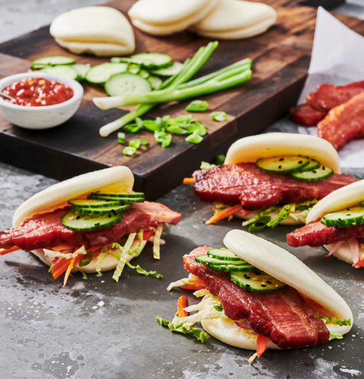 Steamed bao buns with thick-cut bacon, cucumber slices, hoisin sauce, wasabi mayo, sesame seeds, and Asian slaw on a dark slate surface
