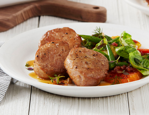 Orange and Thyme Pork Medallions With a Side of Greens on a White Plate and Wooden Table