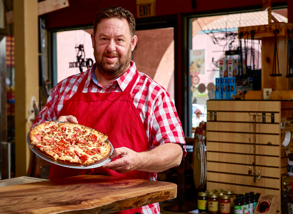 A man in a red apron smiles, holding a pepperoni and sausage pizza