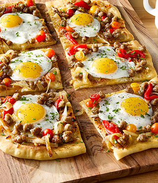 On a wooden board, a pizza topped with sausage, tomatoes, and sunny side up eggs is cut into squares