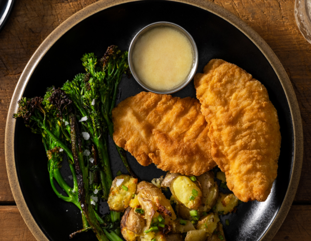 Plate with two breaded steakhouse chicken tenderloins served alongside roasted potatoes, broccolini, and a small cup of sauce