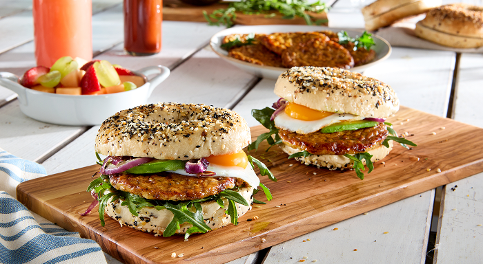 Two breakfast sandwiches on everything bagels with sausage, egg, and avocado on a wooden board