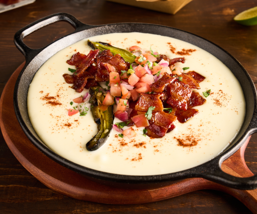 Cast iron skillet of creamy queso topped with hot honey bacon, roasted green chiles, diced tomato and onion, on a wooden table