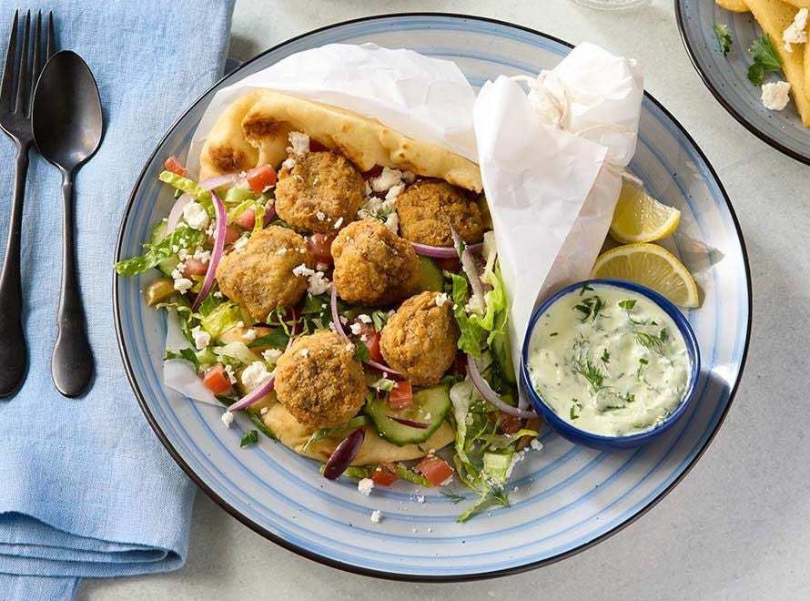 A plate with naan bread wrap stuffed with breaded beef bites, feta cheese and vegetables and a side of lemons and avocado tzatziki