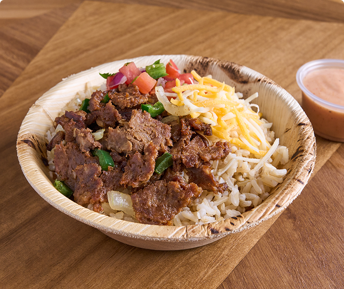 Bowl of sliced steak layered on yellow rice with poblano peppers, melted shredded cheese and ranch sauce