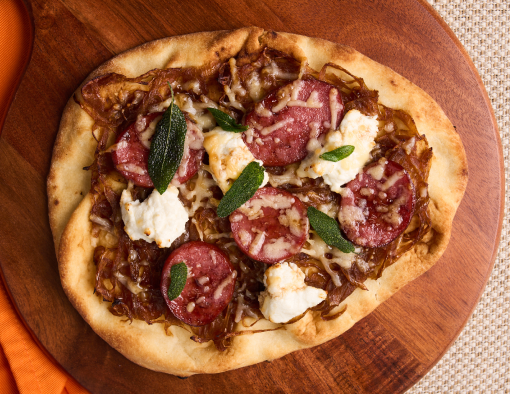 Flatbread with sausage, caramelized onions, ricotta, shredded cheese, and sage on a wooden board