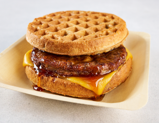 A disposable plate holding a sausage patty sandwiched between two waffles with cheese and maple syrup