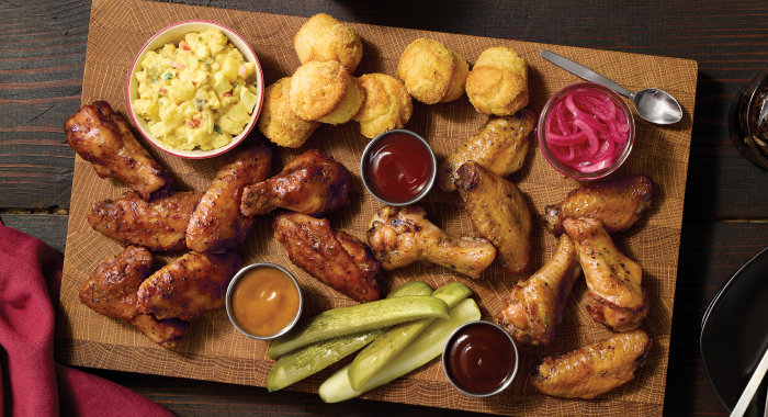 Platter of smoked chicken wings with dipping sauces, pickles, cornbread bites, potato salad, and pickled onions on a rustic wooden board