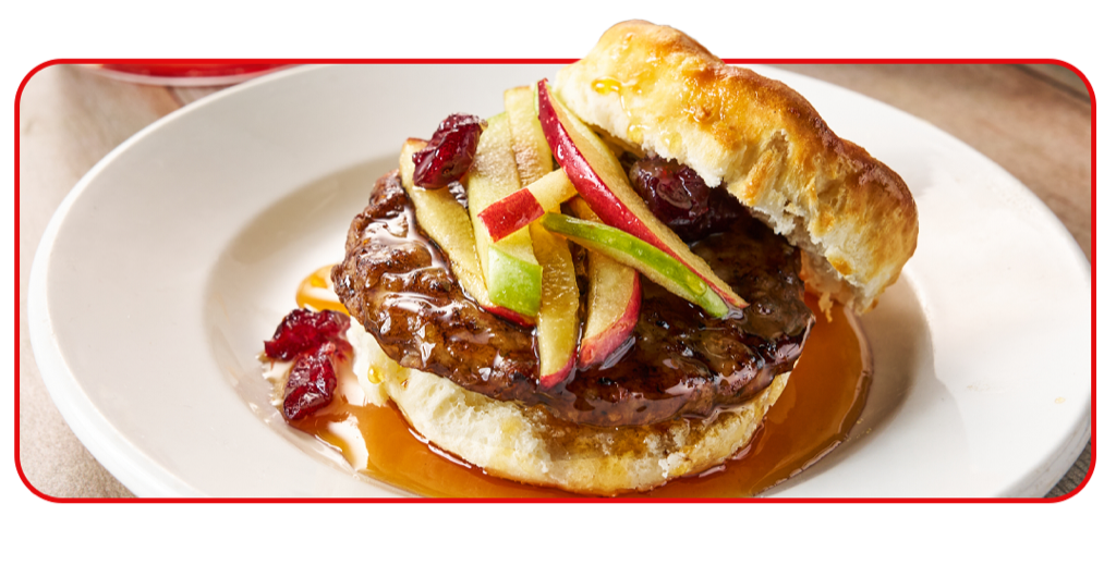 A flaky biscuit open-faced with a pork sausage patty covered in honey and topped with whole cranberries and julienned apples.