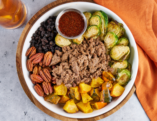 Overhead view of a bowl loaded with sliced beef, acorn squash, pecans, black currants, Brussels sprouts and sauce in a metal ramekin