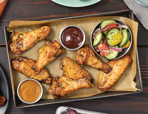 Naturally smoked chicken wings arranged on a parchment-lined tray with barbecue sauce, mustard sauce, and a fresh cucumber tomato salad on the side
