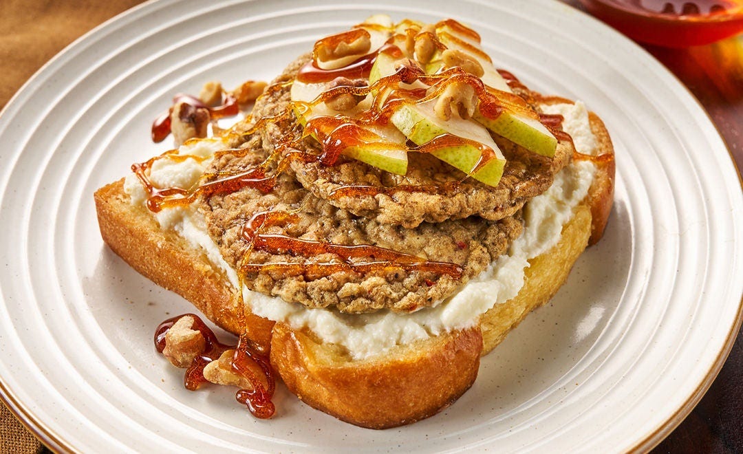 A white plate with thick toast topped with two sausage patties, pear slices and walnuts, drizzled with honey