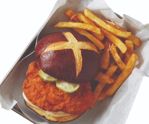 A spicy chicken sandwich on a pretzel bun with pickles that’s placed in a carrier with fries